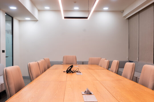 Symmetrical view along an empty boardroom table with beige chairs, a blank white wall for copy space and a conference phone under a glowing pendant frame