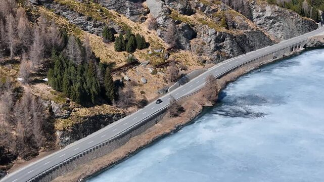 guardrail lined road curves beside frozen silsersee shoreline lone car passes cyclist pedals rocky cliff face towers above larch forest engadin valley switzerland drone aerial view