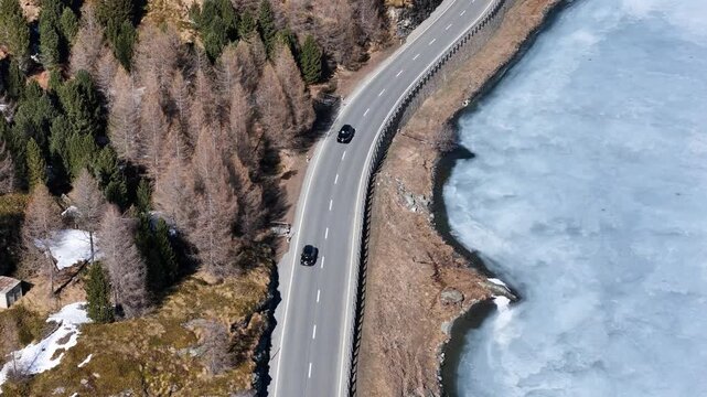 narrow road follows frozen silsersee shoreline single car passes wooden cabin nestled larch forest patchy snow blankets ground engadin valley switzerland drone aerial overhead winter view