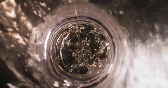 Slow motion pour of carbonated liquid beer into glass bottle from overhead closeup in studio sink setting, probe view.