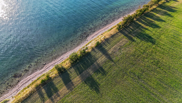 Drohnenaufnahme zeigt ein blaues Gew&auml;sser mit klarem Wasser und Steinufer, eine Baumreihe und ein gr&uuml;nes Feld unter sonnigem Licht, geometrischer Aufbau