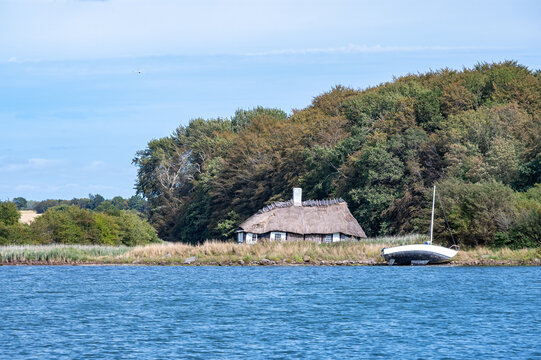 Reetgedecktes Kate-Haus, halb verfallen steht idyllisch an dicht bewaldetem Ufer mit gestrandetem Segelboot am blauen Gew&auml;sser unter sonnigem Himmel.