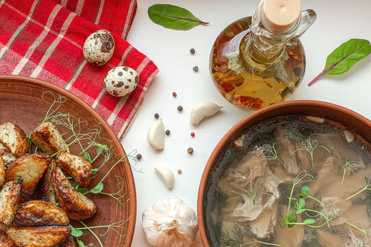Traditional Ukrainian food flat lay with homemade meat kholodets aspic jelly, crispy baked potato wedges with microgreens, quail eggs, garlic cloves, black pepper, and oil in glass bottle.