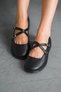 Closeup of female feet in black Mary Jane shoes with crossed straps and metal buckles, standing on textured concrete floor under soft studio lighting