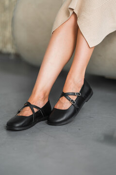 View of lower legs and feet wearing black Mary Jane shoes with crossed straps and shiny metal buckles on textured concrete floor, styled for fashion catalog display under soft light