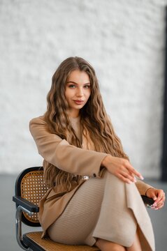 Young woman with long wavy light brown hair in beige blazer and ribbed skirt seated on cane chair, arms crossed and hand reaching toward camera in neutral studio