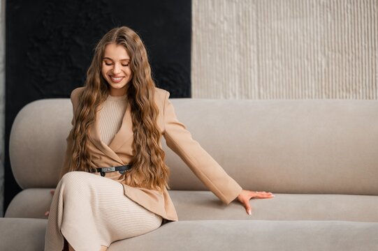Smiling young woman with long wavy hair seated on modern neutral sofa wearing beige blazer and ribbed knit dress