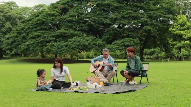 GrandFather playing acoustic guitar for child during picnic. Asian family enjoy outdoor picnic with food and drinks in park sunny day, outdoor activitie with daughter father, mother grandfather