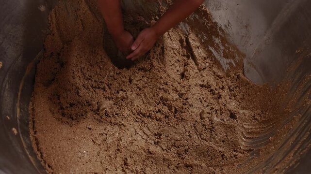 Hands work dense, dark rye bread dough inside a large wooden vessel, showing the rustic texture and traditional process of bread making