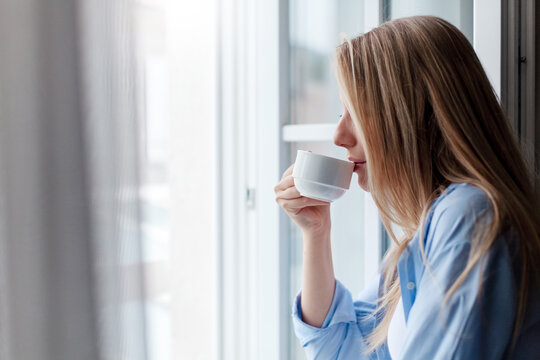 Young woman drinking coffee by opened window. Happy girl relaxing, enjoying morning routine at home. Concept of slow living, mindfulness, female mental health