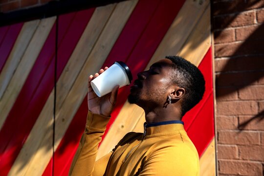 Person in a yellow jacket enjoying a drink outdoors by a red wall.