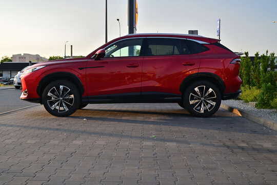 New generation red MG HS SUV parked on a city cobblestone parking lot, side view.