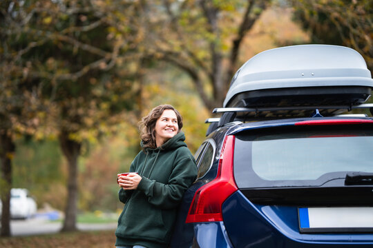 A smiling woman in a green hoodie leaning against a blue car with a roof cargo box while holding a hot mug outdoors during autumn