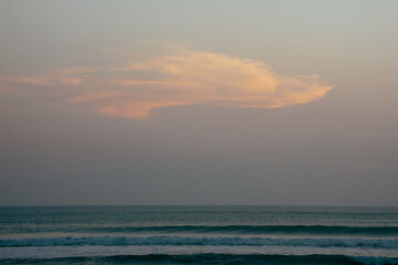 Fototapeta premium A serene sunset over the ocean in Bali features a large, orange cloud against a pastel sky, with gentle waves lapping at the shore.