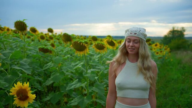 Woman walks through sunflower field at sunset. She wears white outfit and headband. Her gaze is downcast, showing calmness and reflection. Golden sunflowers stretch to horizon under soft sky