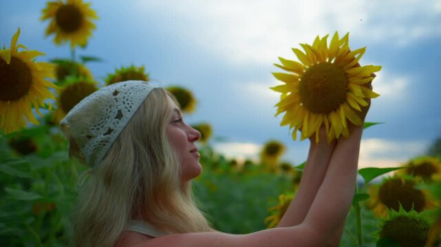 Woman smiles while holding a sunflower. She wears a light knit cap. Sunflowers stretch toward the sky. Sky shows clouds against soft daylight. Footage fits summer, nature, or happiness themes