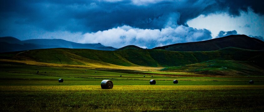 Dramatic storm clouds over scenic rolling green hills and grassy field with round hay bales in beautiful rural landscape under dark blue sky