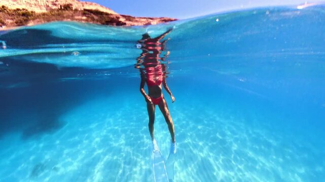 Freediver in red bikini and long fins swimming in turquoise ocean. Over under split shot.