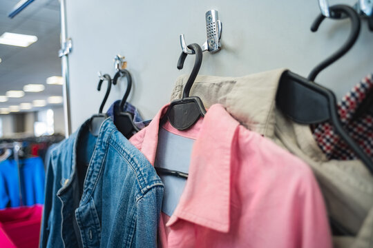 Close-up view of secondhand clothing hanging on plastic hangers inside retail store aisle, featuring denim jacket, pink blouse, beige coat, and patterned garment under bright fluorescent lighting