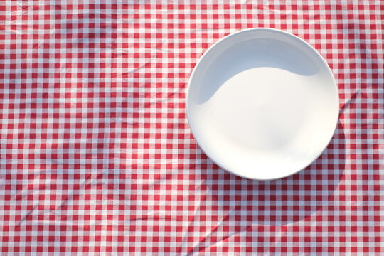 An empty white plate on a red checkered tablecloth, viewed from above. Flat lay. Kitchen mockup.