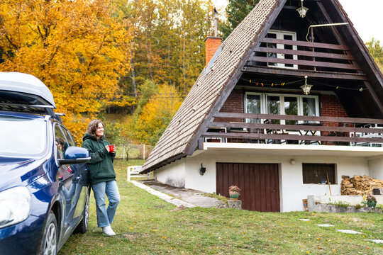 A woman leaning against a blue car with a red mug in her hand in front of a wooden A-frame house surrounded by colorful autumn forest