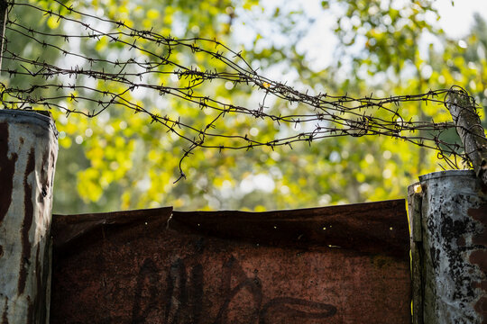 Rusted barbed wire stretches between two weathered cylindrical metal posts above a torn corrugated sheet metal panel, forming an improvised rural perimeter fence