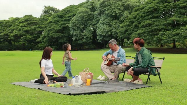 GrandFather playing acoustic guitar for child during picnic. Asian family enjoy outdoor picnic with food and drinks in park sunny day, outdoor activitie with daughter father, mother grandfather