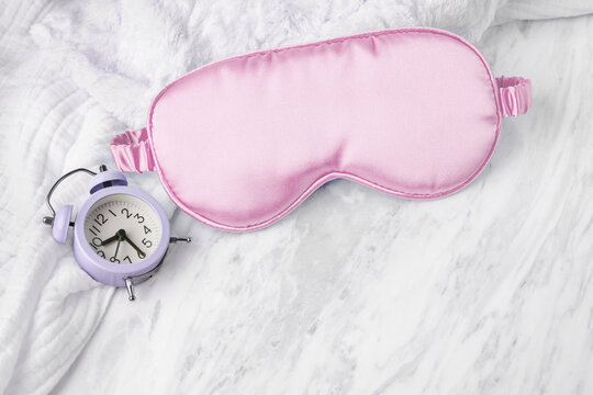Pink sleep mask and alarm clock on soft towel on white marble background