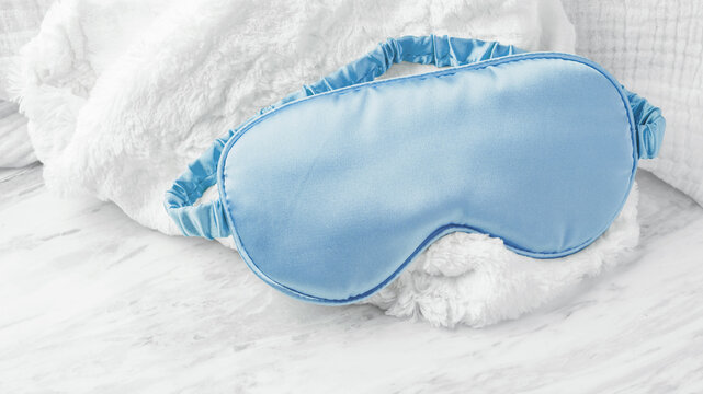 Blue sleep mask on soft fabric on marble background, closeup