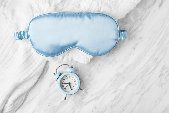 Sleep mask and alarm clock on soft towel on white marble background, copy space
