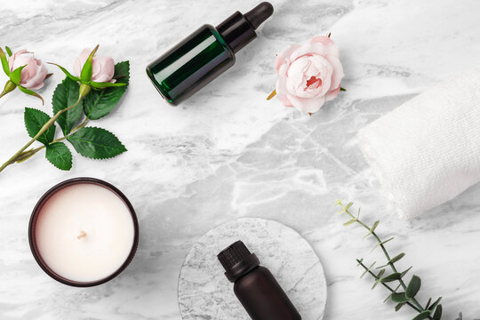 Flat lay of essential oils, scented candle, rose flowers, and serum bottle on white marble background