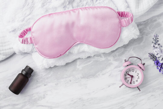 Silk sleep mask on soft fabric with essential oil and alarm clock on marble background, top view