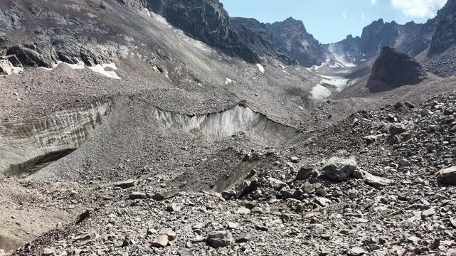 Cinematic drone flyover over rocky peaks, Oktyabrskaya cave, and shrinking ice blocks near Talgar Pass at 3400 meters altitude, revealing climate change and glacier melting in Tian Shan mountains near