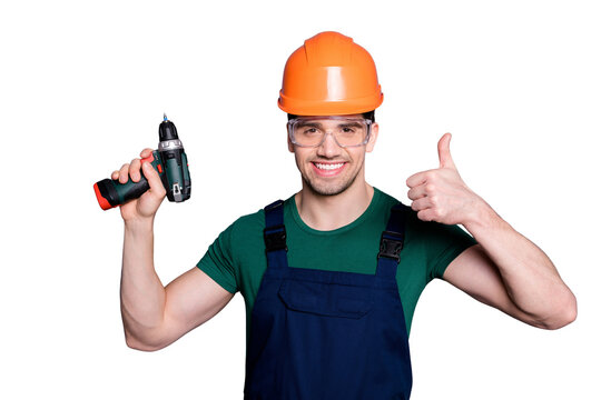 Portrait of handsome man repairer successful handyman hold perforator show thumb up sign wear orange hard hat green t-shirt protective goggles isolated over grey color background