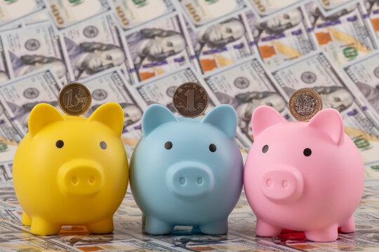 Three piggy banks containing 1 Chinese yuan, 1 British pound, and 1 euro coins, set against a background of 100 US dollar banknotes.