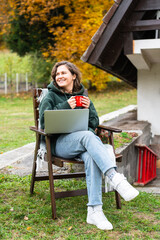 Obraz premium A woman sitting in a wooden chair outside an A-frame house with a laptop and a red mug during autumn