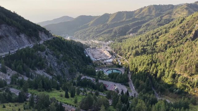 Cinematic drone flyover moving from the crest of Medeu dam toward mountain gorge with winding serpentine road, ice rink, and Almaty city panorama under golden sunset sky in Kazakhstan at summer