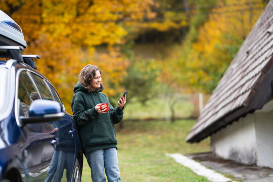 A smiling woman standing next to a blue car with a roof box, holding a red mug and using a smartphone in front of an A-frame house in autumn