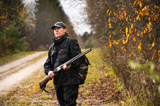 A hunter in camouflage gear with a backpack and rifle walking along a dirt road through an autumn forest