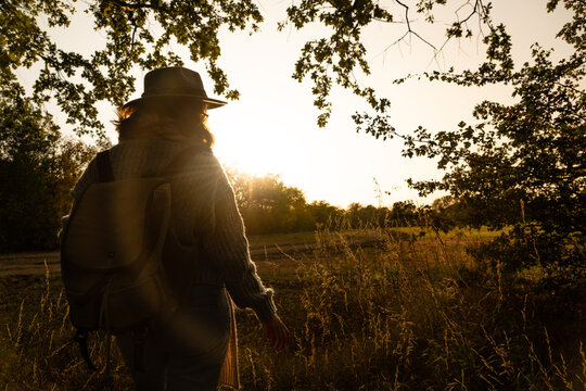 A woman in a hat and sweater with a backpack standing in a grassy field looking towards the warm golden sunset