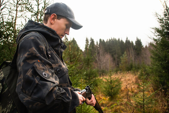 A hunter in a cap and camouflage jacket inspecting his shotgun while outdoors in an autumn forest edge