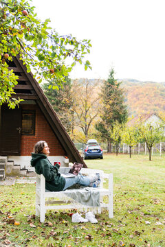 A person sitting outdoors on a wooden bench with a laptop during autumn in front of a rural house and a car.