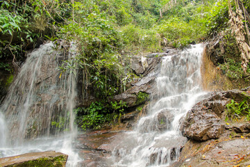 Tropical jungle waterfall in Thailand surrounded by lush green rainforest, moss-covered rocks, dense vegetation, and flowing water. The natural cascade creates a peaceful and refreshing scene © Yuliia