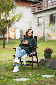 A woman sitting in a wooden chair with a blanket and a red mug, relaxing outdoors during autumn in a countryside garden