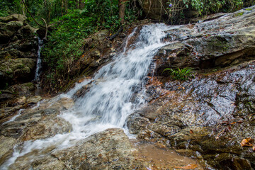 Tropical jungle waterfall in Thailand surrounded by lush green rainforest, moss-covered rocks, dense vegetation, and flowing water. The natural cascade creates a peaceful and refreshing scene © Yuliia