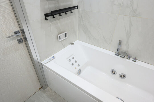 Modern compact bathroom features white jacuzzi tub with jets, black wall shelf, marble tiles, chrome fixtures, handheld showerhead, control panel, minimalist design