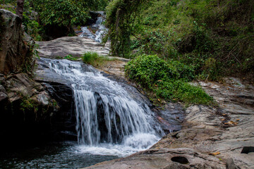 Tropical jungle waterfall in Thailand surrounded by lush green rainforest, moss-covered rocks, dense vegetation, and flowing water. The natural cascade creates a peaceful and refreshing scene © Yuliia