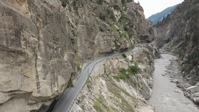 Aerial video of a yellow vehicle driving on a road clinging to a sheer vertical cliff face hundreds of metres above a fast-flowing grey river in a deep canyon in Spiti Valley Himachal Pradesh