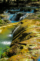 Tropical jungle waterfall in Thailand surrounded by lush green rainforest, moss-covered rocks, dense vegetation, and flowing water. The natural cascade creates a peaceful and refreshing scene © Yuliia