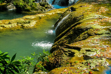 Tropical jungle waterfall in Thailand surrounded by lush green rainforest, moss-covered rocks, dense vegetation, and flowing water. The natural cascade creates a peaceful and refreshing scene © Yuliia